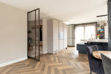 Moderne stalen scheidingswand en deuren in een open woonkeuken met houten vloer en grote ramen voor natuurlijk licht.