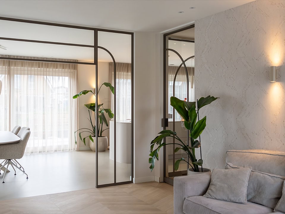 Luxe interieur met stalen deuren en grote ramen, natuurlijke lichtinval, groene planten en comfortabele zithoek, perfect voor moderne woninginrichting.