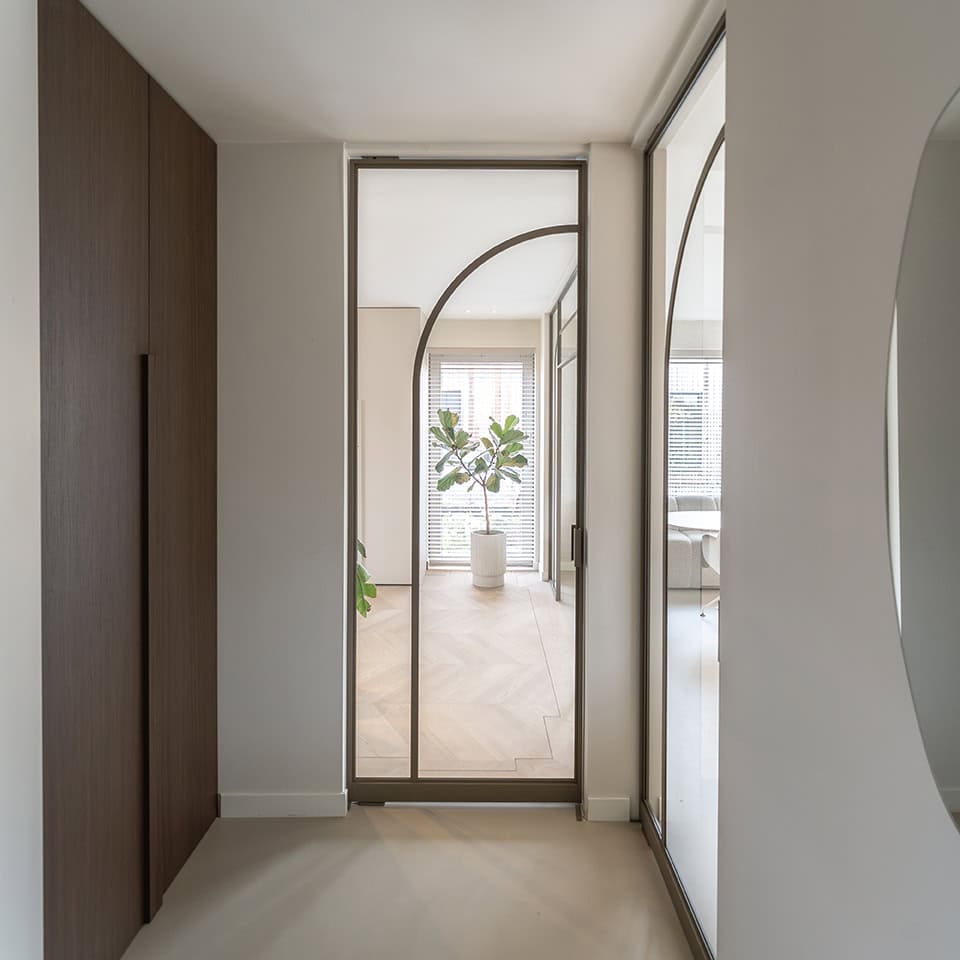 Moderne stalen deur met glas en houten vloer, scheidt twee ruimtes in een eigentijdse woning, met natuurlijke lichtinval en planten op de achtergrond.