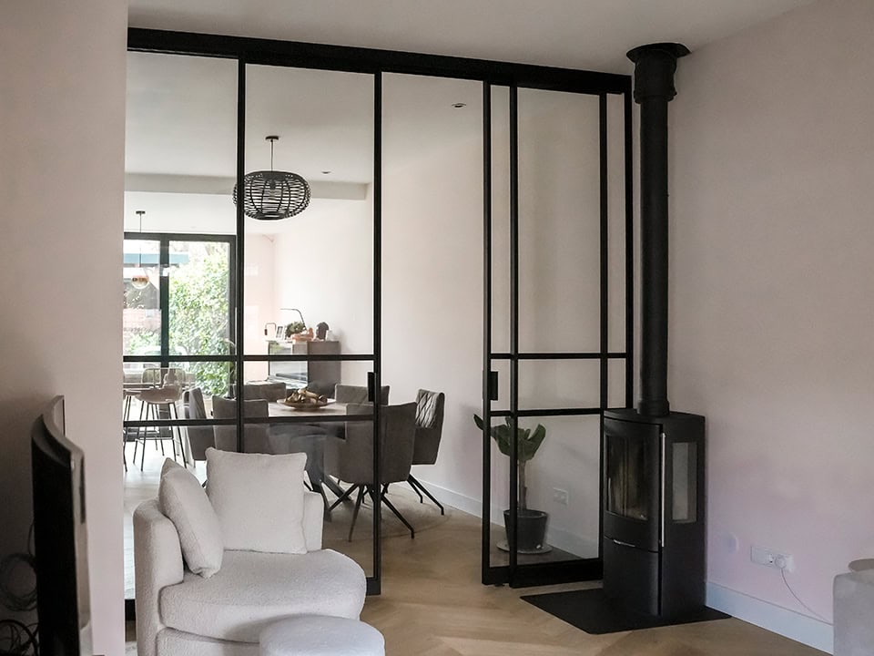 Strakke stalen scheidingswand met glas in een moderne woonkamer, scheidt de eet- en zithoek, met een houtkachel en natuurlijke lichtinval.