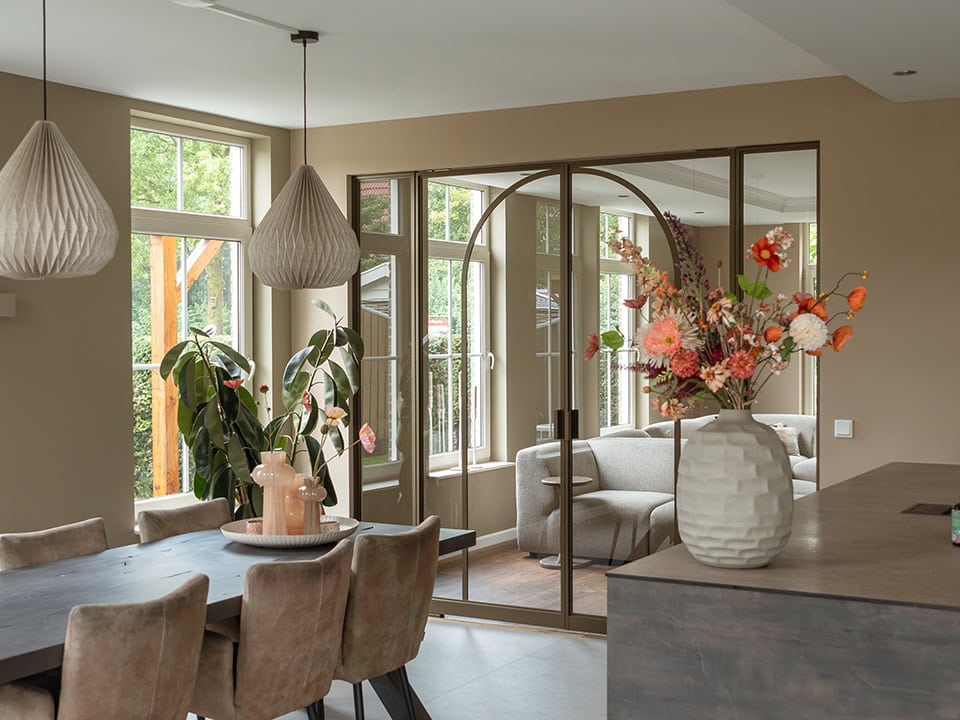 Lichte woonkamer met grote ramen, moderne stalen scheidingswand en decoratieve bloemen in een witte vaas, ideaal voor stijlvolle interieurs.