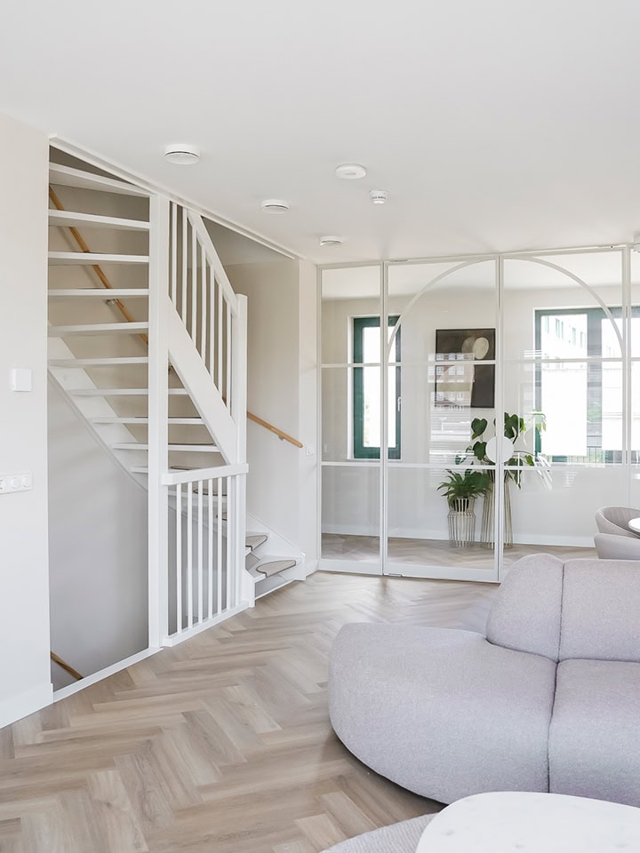 Lichte en moderne woonkamer met glazen scheidingswand, witte trap en comfortabele bank, perfect voor een stijlvolle woninginrichting.