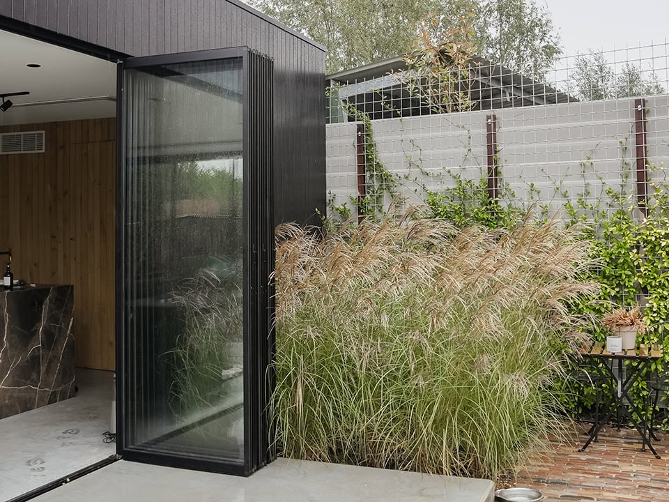 Moderne opvouwbare stalen scheidingswand met glazen panelen, ideaal voor binnen- en buitengebruik, met een tuin en natuurlijke elementen op de achtergrond.