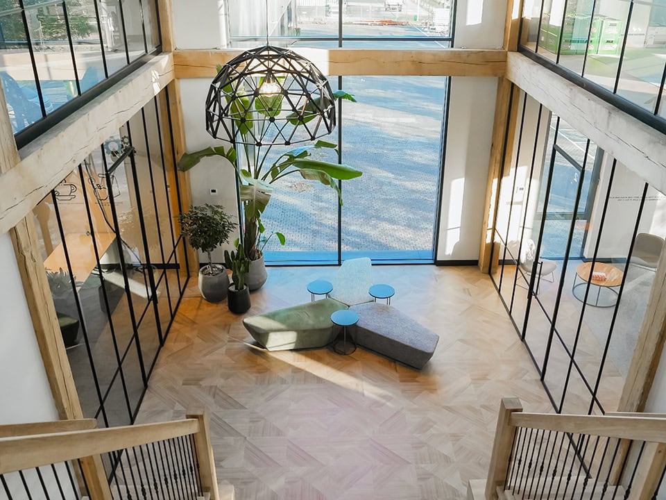 Lichte kantoorruimte met grote glazen wanden, houten vloeren en moderne meubels, inclusief een plantenwand en zitgelegenheid, geschikt voor een professionele werkomgeving.