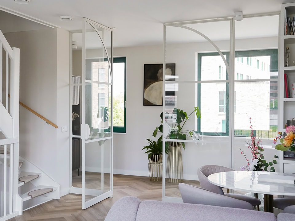 Lichte en moderne stalen scheidingswanden in een woonkamer met grote ramen, planten en decoratieve elementen, passend bij hoogwaardige stalen deuren en wanden op maat.
