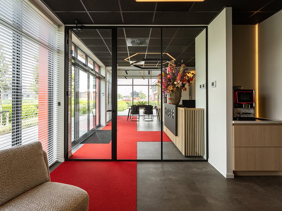 Binnenkant van een kantoor met een glazen stalen scheidingswand, grote ramen, een rode tapijt en een bloemenarrangement, passend bij de focus op stalen deuren en wanden.