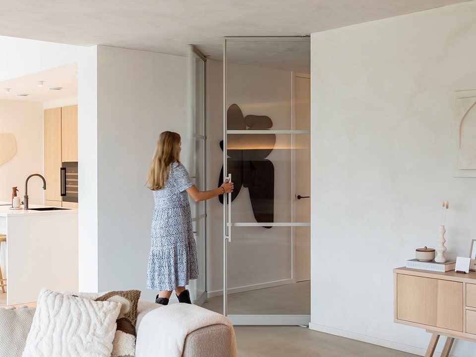 Een jonge vrouw opent een glazen schuifdeur in een modern, minimalistisch interieur met witte muren en houten meubels, passend bij hoogwaardige stalen deuren en wanden.
