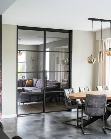 Moderne stalen schuifdeur met glazen panelen scheidt woonkamer van eetkamer, industrieel design, zwart frame, natuurlijke lichtinval, stijlvolle interieurafwerking.