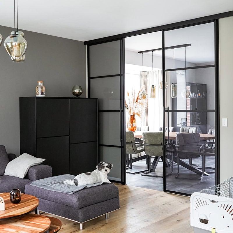 Moderne stalen schuifdeur met zwart frame en glazen panelen, scheidt woonkamer en eetkamer, geeft een industriële uitstraling aan het interieur.