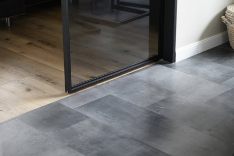 Moderne stalen schuifdeur met glazen panelen, scheidt woonkamer van keuken, met grijze tegelvloer en houten vloer in achtergrond.