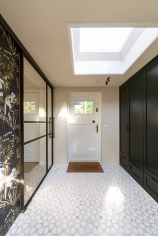 Moderne entree met grote stalen schuifdeuren, natuurlijke lichtinval via dakraam, zwarte op maat gemaakte kast, en decoratieve wand met botanisch patroon.